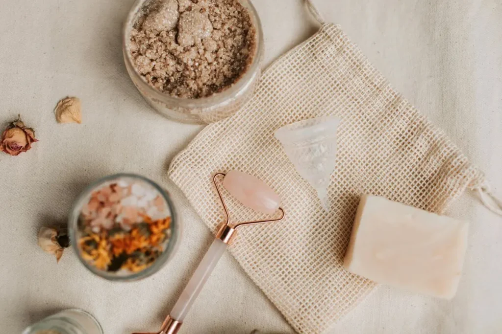 Flat lay de autocuidado natural com roller facial de quartzo rosa, sabonete artesanal, sais de banho, flores secas e tecido de algodão em tons neutros.
