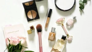 Flat lay de produtos de beleza sobre fundo claro, com maquiagem, pincel, perfume, roller facial, caderno e flores rosas em composição delicada.