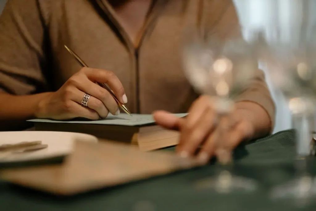 Mulher escrevendo em um caderno com caneta, em ambiente acolhedor e luz suave, representando reflexão, organização emocional e autocuidado.