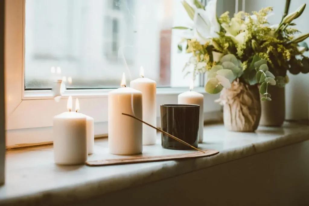 Velas brancas acesas sobre a janela, com incenso, vaso de flores e luz natural suave, transmitindo calma, aconchego e bem-estar.
